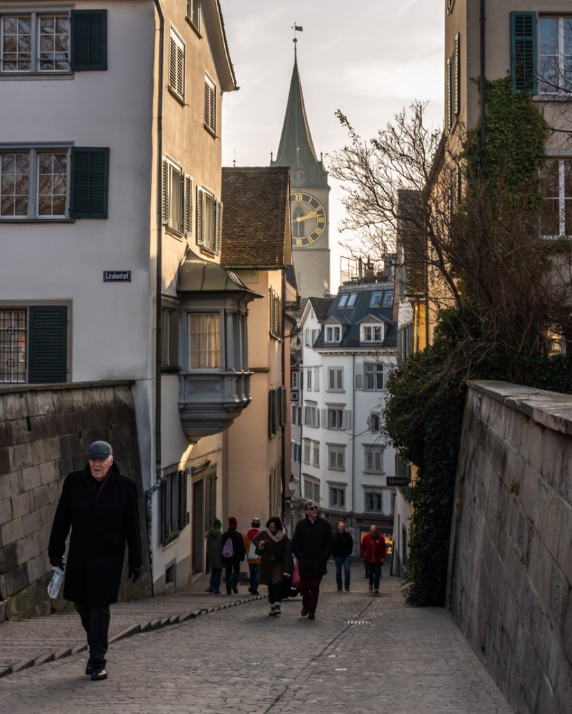 People walk up a cobbled lane in Zurich with a church spire in the background.