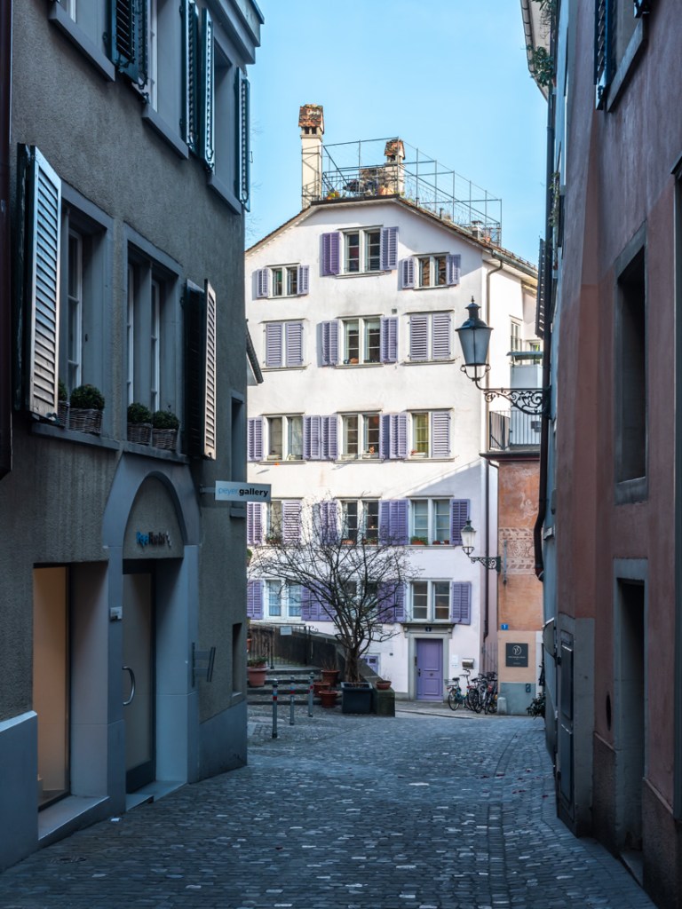 A cobbled alleyway in Zurich with a historic building with magenta window shutters.